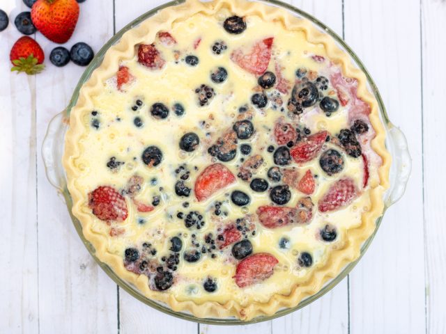 an overhead shot of a whole pie with blueberries, blackberries and strawberries that has a creamy custard filling poured inside the pie plate. There’s fresh berries in the background.