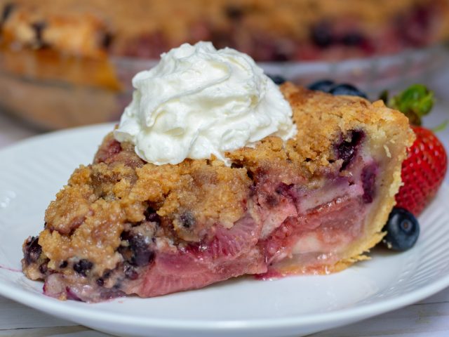 a slice of berry pie on a small white plate with blackberries, blueberries and strawberries. It’s topped with fresh whipped cream and there’s fresh berries on the plate.