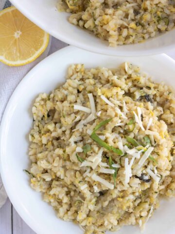 A white bowl filled with creamy summer squash risotto. It's topped with fresh chopped basil and shredded cheese. There's a slice of lemon in the background and a big serving bowl with risotto.