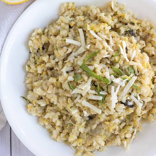 A white bowl filled with creamy summer squash risotto. It's topped with fresh chopped basil and shredded cheese. There's a slice of lemon in the background and a big serving bowl with risotto.