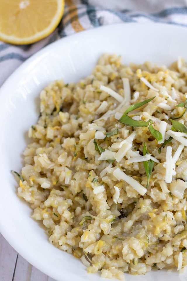 A white bowl filled with creamy summer squash risotto. It's topped with fresh chopped basil and shredded cheese. There's a slice of lemon in the background and a yellow and navy blue plaid napkin.