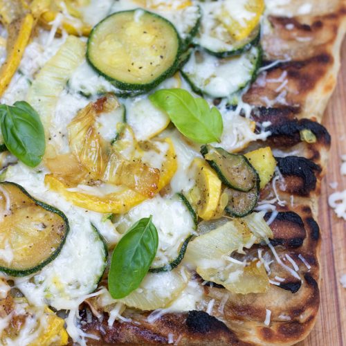Grilled pizza on a wooden cutting board with shredded cheese sprinkled on the board. The crust has slight char marks on it and it's topped with summer squash, onions and fresh basil