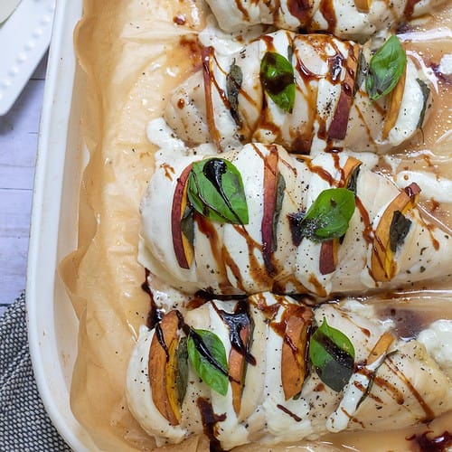 A white baking dish lined with brown parchment paper with 4 pieces of hasselback peach caprese chicken breasts that have been baked in the oven. They're drizzled with balsamic and have fresh basil and melted mozzarella. There's a small dish of fresh basil and peach slices in the background.