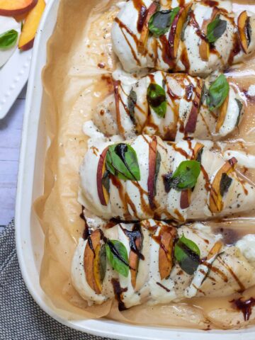 A white baking dish lined with brown parchment paper with 4 pieces of hasselback peach caprese chicken breasts that have been baked in the oven. They're drizzled with balsamic and have fresh basil and melted mozzarella. There's a small dish of fresh basil and peach slices in the background.