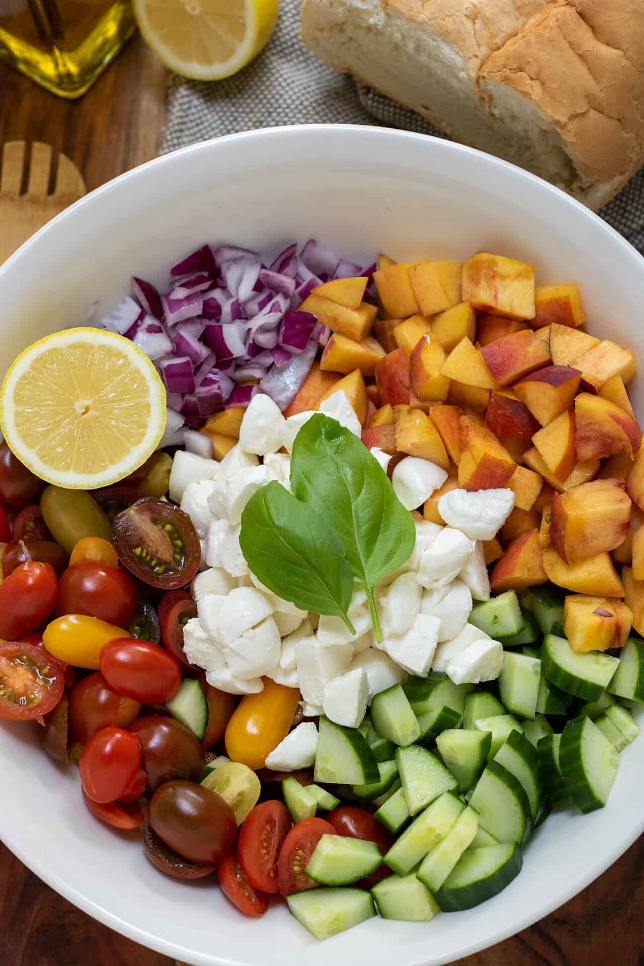 A large white bowl with halved cherry tomatoes, diced cucumber, red onion, diced peaches, a half a lemon, & fresh basil leaves. There's a loaf of Italian bread in the background with a wooden serving spoon and olive oil