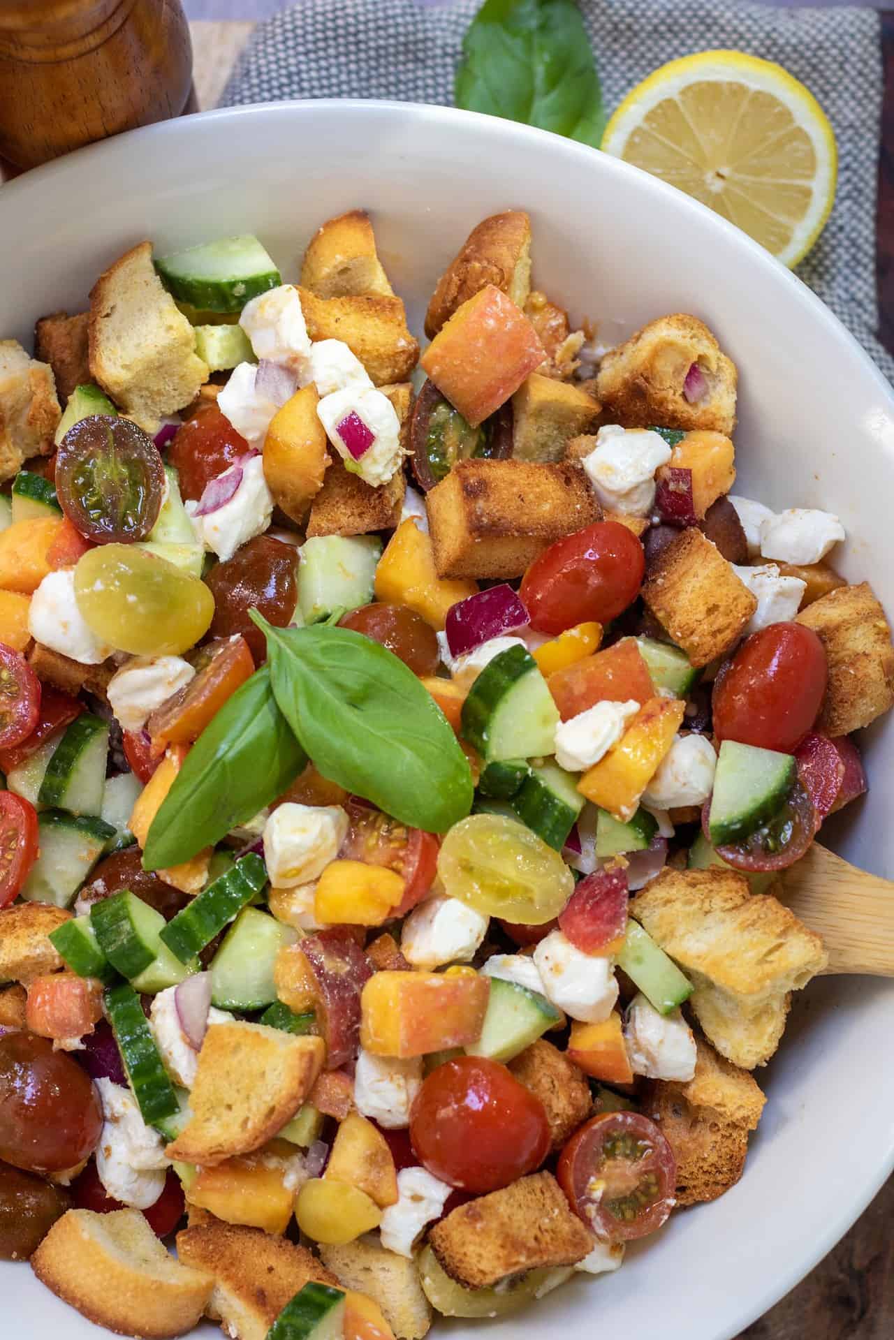 A large white bowl with chunks of golden crispy bread, cherry tomatoes, cucumbers, red onion slices, fresh basil, mozzarella and peaches.