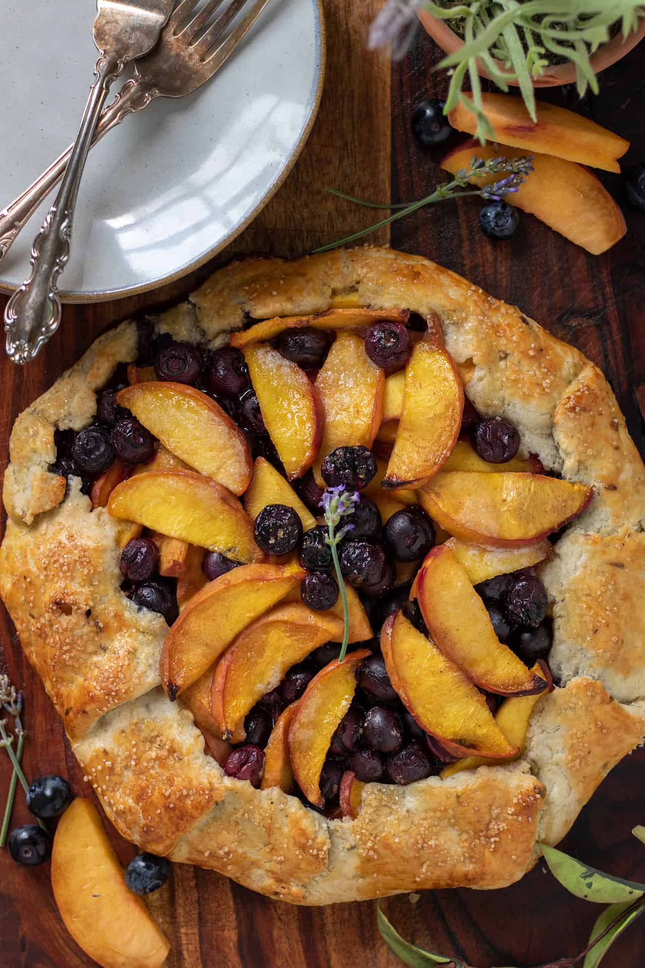 Peach blueberry crostata on top of a wooden background. There's a stack of small dessert plates in the corner with two forks. There's peach slices and blueberries spread on the wooden board. There's a sprig of fresh lavender in the middle of the baked crostata.