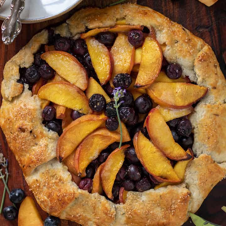 Peach blueberry crostata on top of a wooden background. There's a stack of small dessert plates in the corner with two forks. There's peach slices and blueberries spread on the wooden board. There's a sprig of fresh lavender in the middle of the baked crostata.