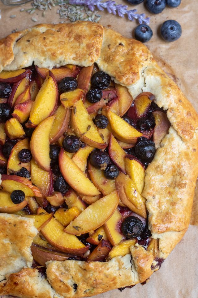 A peach blueberry crostata that's been baked on parchment paper. The crust is golden brown. There's a couple of fresh blueberries with dried lavender and a purple lavender flower in the background.