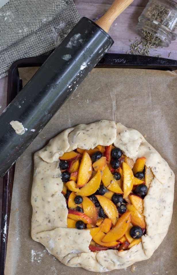 A baking sheet lined with parchment paper with a peach blueberry crostata ready to be baked. There's a rolling pin with flour on it and a small jar of dried lavender spilled in the background.
