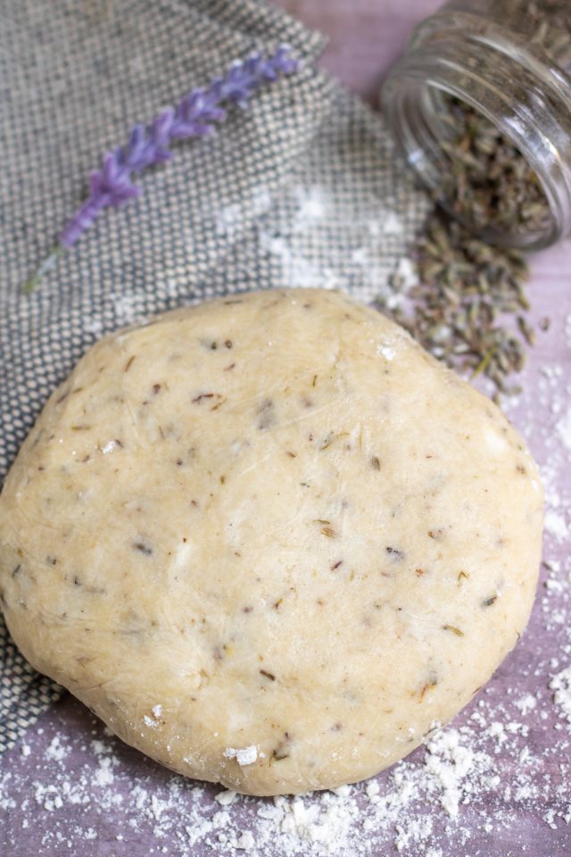 A disc of crostata dough that's laced with dried lavender. The surface is sprinkled with flour and there's a kitchen towel, small jar of dried lavender, and a fresh lavender flower in the background.
