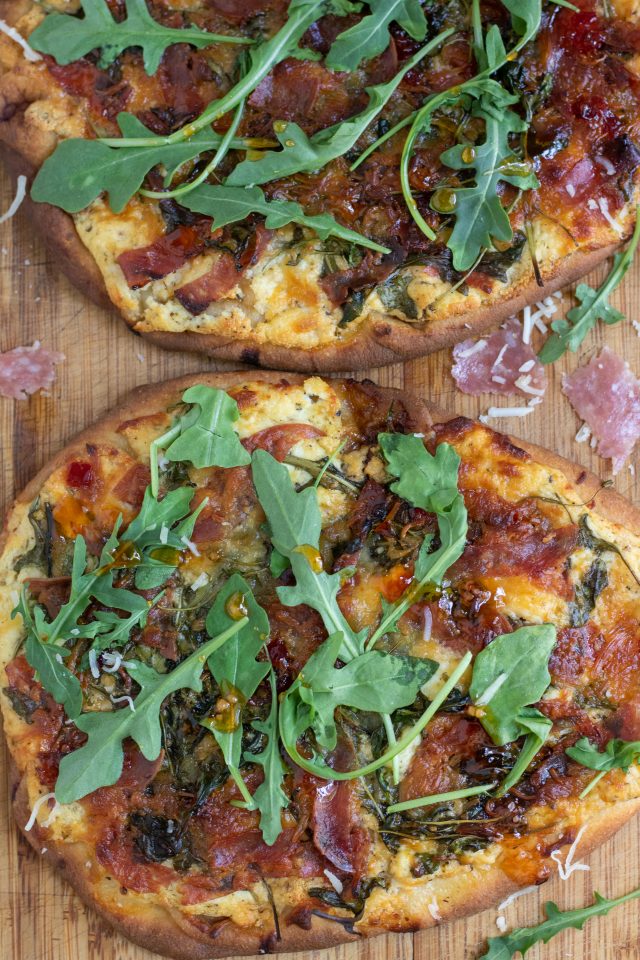 A wooden cutting board with 2 flatbread pizzas that are crispy and golden brown. They're topped with slices of salami, ricotta and goat cheese, arugula and drizzled with red pepper jelly. There's shredded cheese and arugula sprinkled on the cutting board and slices of fresh salami.