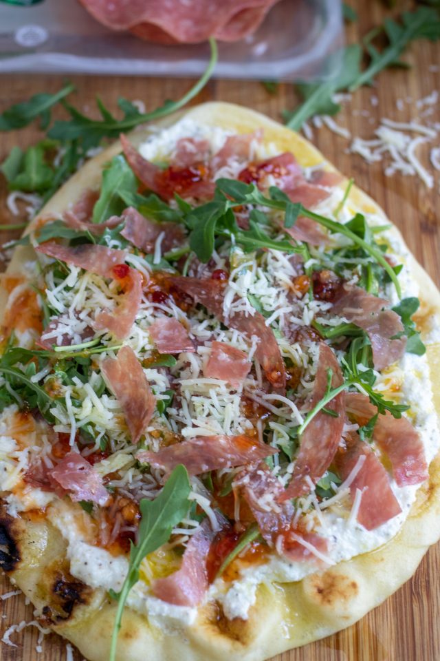 A wooden cutting board with a nann flatbread pizza that's topped with cheese, arugula and slices of salami. It's drizzled with red pepper jelly and you can see shredded cheese sprinkled on the cutting board and slices of salami in the background.