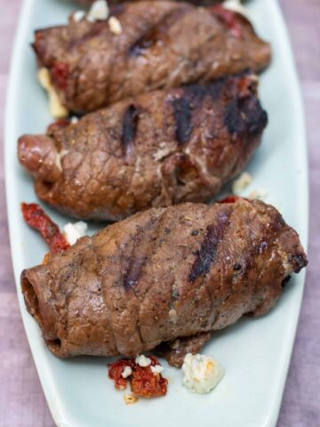 a light blue, oblong serving platter with 3 steak roll ups that have slight char marks and are browned. There’s some gorgonzola crumbles on the platter and some sun dried tomatoes.