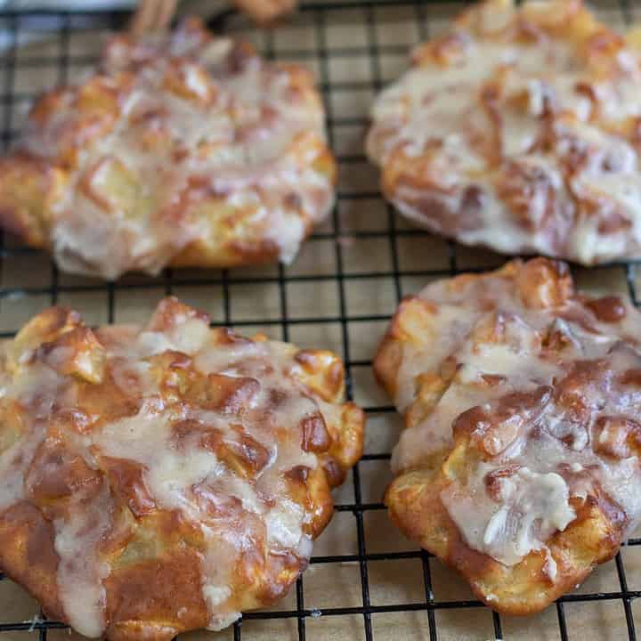 A cooling rack with brown parchment underneath it with air fried apple fritters on top. They're golden brown and have glaze dripping down the sides. There's a cup of coffee in the background with a red apple and two whole cinnamon sticks.