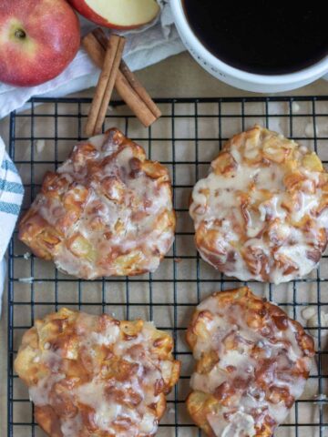 Air fried apple fritters on top of a cooling rack with parchment paper. They're dripping with brown butter glaze. There's a cup of coffee and red apple in the background with two whole cinnamon sticks.