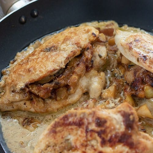 A large saute pan with stuffed chicken breasts that are in a brandy cream sauce. There's an apple in the background and bottle of brandy.