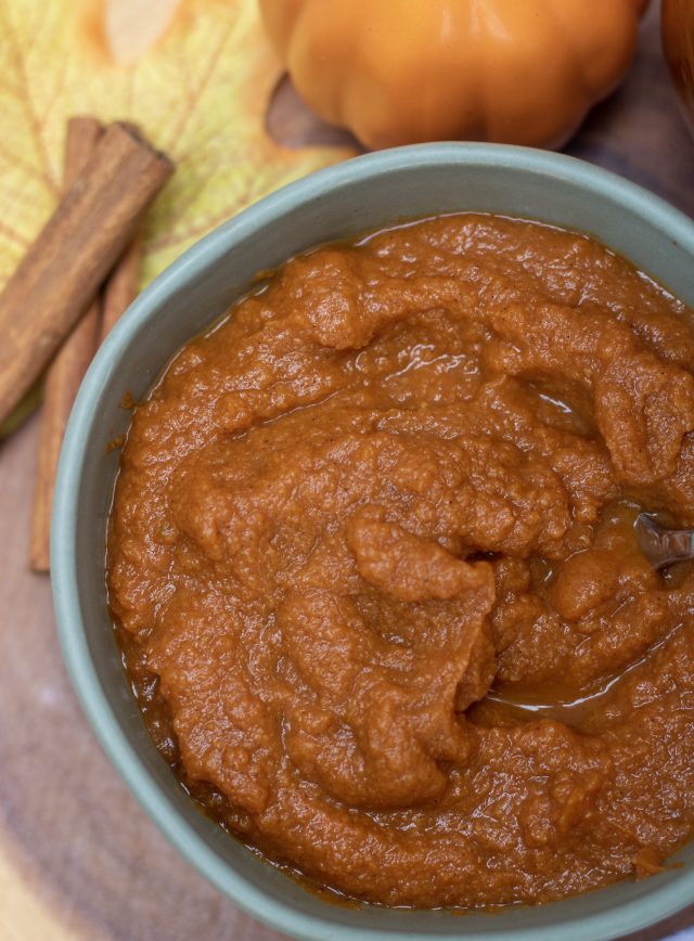 An overhead closeup shot of easy homemade pumpkin butter in a small green bowl. There’s a cinnamon stick and yellow leaf in the background
