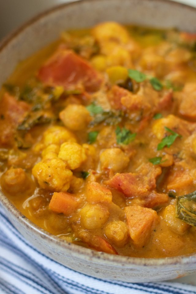 A bowl full of creamy pumpkin coconut curry made with chickpeas and vegetables. There's a blue and white dish towel next to the bowl. It's sprinkled with fresh cilantro.