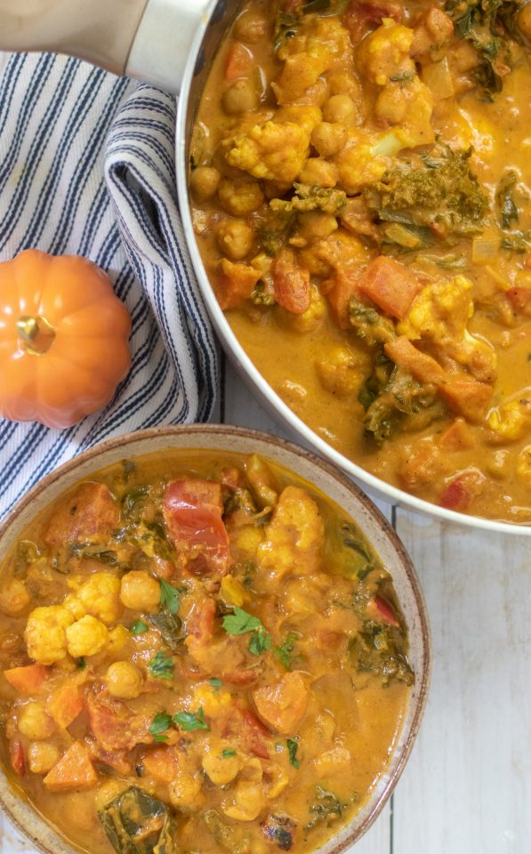 A bowl of healthy and vegan pumpkin coconut curry. There's chickpeas and cauliflower in it with a sprinkle of fresh cilantro. A large pot of the curry is in the background with a small orange pumpkin.