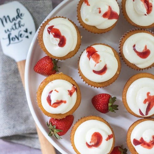 An overhead shot of roasted strawberry-filled cupcakes on top of a white cake stand. There's strawberry reduction drizzled over the tops of the cupcakes with fresh whole strawberries scattered on the plate. There's a spatula next to it that says "made with love" with a bunch of fresh strawberries next to it.