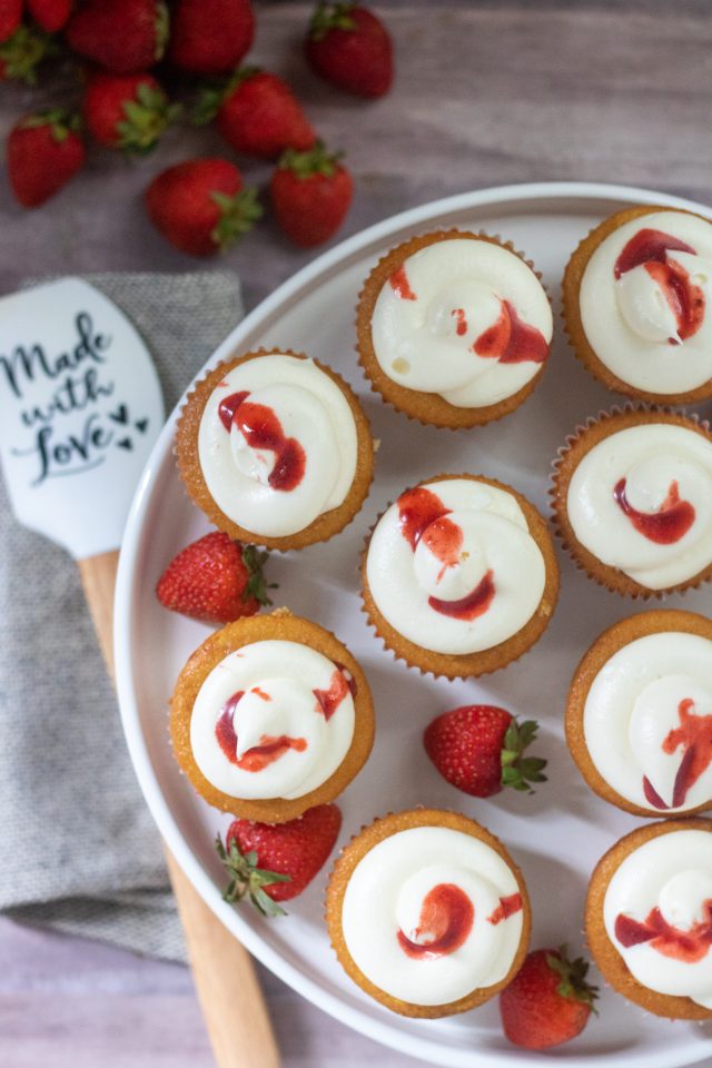 An overhead shot of roasted strawberry-filled cupcakes on top of a white cake stand. There's strawberry reduction drizzled over the tops of the cupcakes with fresh whole strawberries scattered on the plate. There's a spatula next to it that says "made with love" with a bunch of fresh strawberries next to it.