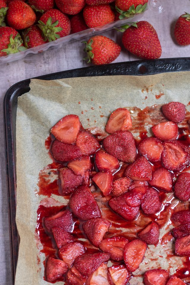 A sheet pan lined with brown parchment paper with juicy roasted strawberries on top of it. A carton of fresh strawberries is next to the pan.