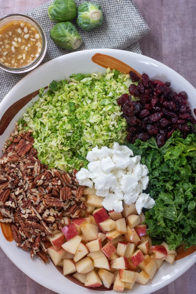 A large bowl with shredded brussels sprouts, chopped kale, chopped toasted pecans, goat cheese crumbles, diced apples and dried cranberries. There's a mason jar of dressing next to the bowl with 3 whole brussels sprouts.