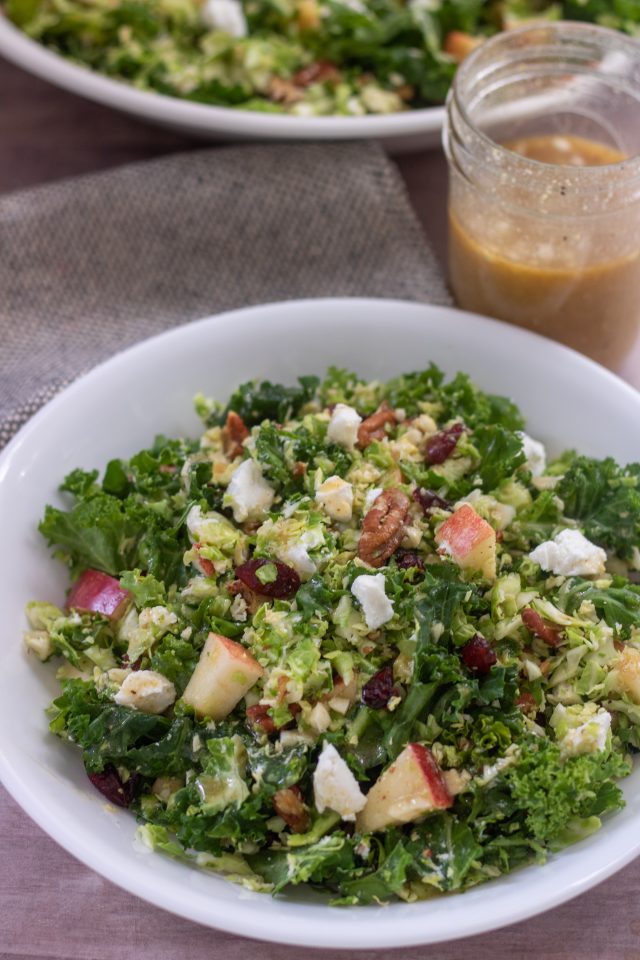 A wide white bowl filled with shredded brussel sprout salad with cranberries, goat cheese, apples, pecans and kale. It's topped with maple dijon dressing. There's a small mason jar of dressing behind the bowl with a glimpse of a bigger serving bowl full of the fall salad.