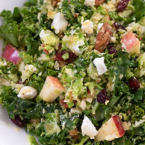 An up close shot of shredded brussels sprout and kale salad. It's topped with toasted pecans, goat cheese and apples and tossed with a maple dijon dressing.
