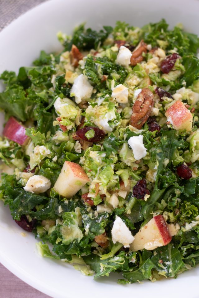 An up close shot of shredded brussels sprout and kale salad. It's topped with toasted pecans, goat cheese and apples and tossed with a maple dijon dressing.