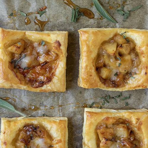A sheetpan lined with parchment paper with golden puff pastry square appetizers filled with apples and cheddar cheese. There's drizzled maple syrup on the pan and it's sprinkled with fresh sage.