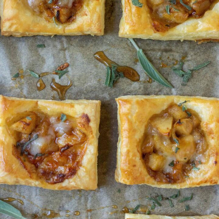 A sheetpan lined with parchment paper with golden puff pastry square appetizers filled with apples and cheddar cheese. There's drizzled maple syrup on the pan and it's sprinkled with fresh sage.