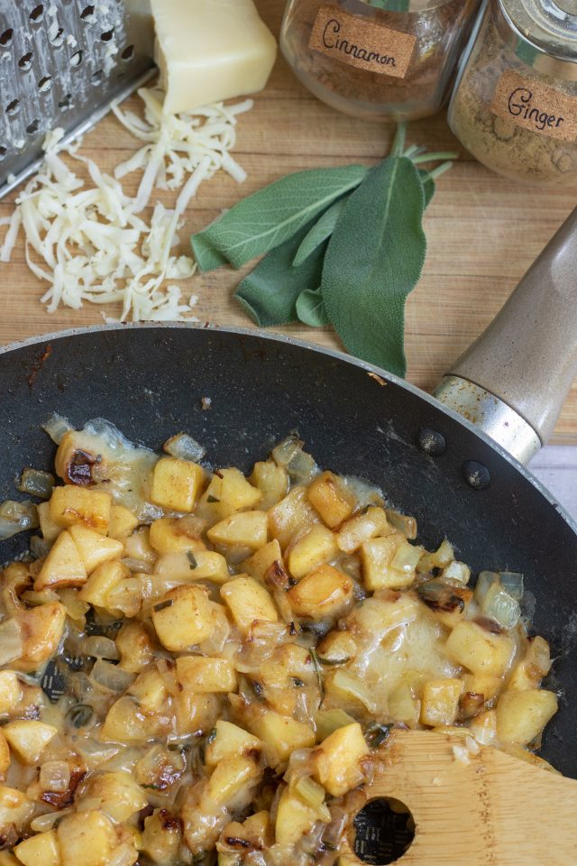 A large saute pan with diced apples, onions and melted cheddar cheese. There's a wooden spoon in the pan. A metal cheese grater with a block of half grated cheese is in the background with a bunch of fresh sage leaves and a jar of cinnamon and ginger spices.