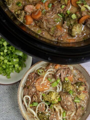 A brown speckled bowl of noodles with asian beef, carrots, broccolis and bell pepper. It's topped with sesame seeds and scallions. There's a small bowl full of scallions next to it and a black crockpot filled with the thai beef stew in the background.