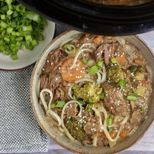 A brown speckled bowl of noodles with asian beef, carrots, broccolis and bell pepper. It's topped with sesame seeds and scallions. There's a small bowl full of scallions next to it and a black crockpot filled with the thai beef stew in the background.