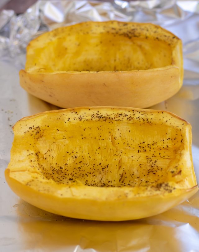 A foil lined sheet pan with 2 halved spaghetti squash that's sprinkled with salt and pepper and has been baked until tender.