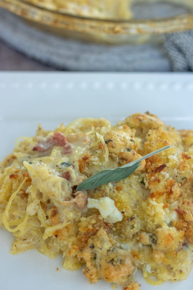 A white square plate with a serving of creamy spaghetti squash gratin on it. You can see the small pancetta peices and gorgonzola crumbles. It's topped with a sage leaf.