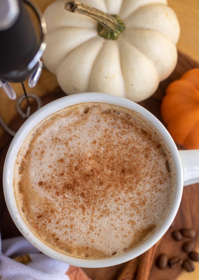 A white coffee mug with pumpkin latte sprinkled with cinnamon. It's on a wooden circle with a small white and orange pumpkin