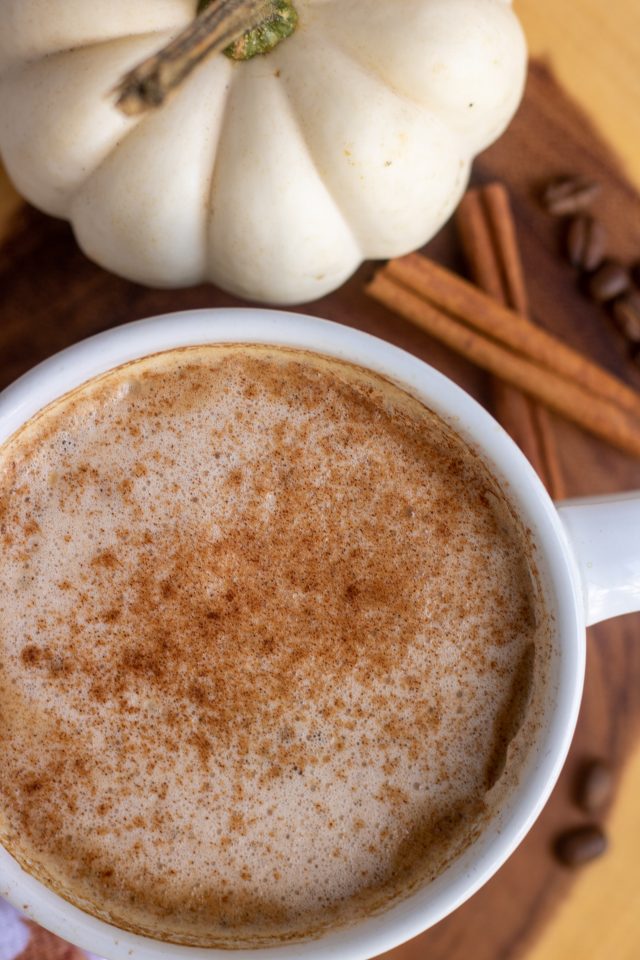 A white coffee mug with pumpkin latte sprinkled with cinnamon. It's on a wooden circle with whole cinnamon sticks, coffee beans and a white pumpkin