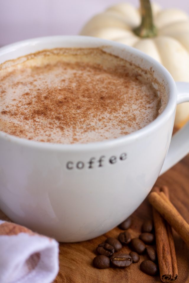 A large white coffee mug that says "coffee" filled with pumpkin spice latte and sprinkled with cinnamon. There's cinnamon sticks and coffee beans in the background with a small white pumpkin