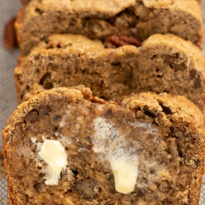 A sliced loaf of pecan cinnamon banana bread. There's butter that's a little melted smeared on the first slice. There's crumbs and whole pecans on the linen cloth.