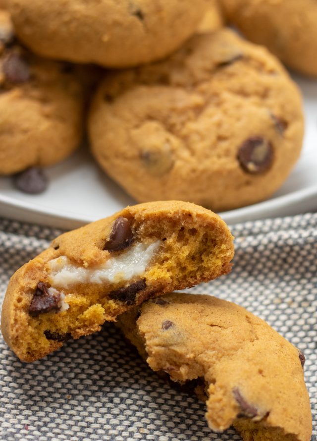 A pumpkin chocolate chip cookie broken in half where you can see the cream cheese filling. There's a beige plate of cookies in the background.