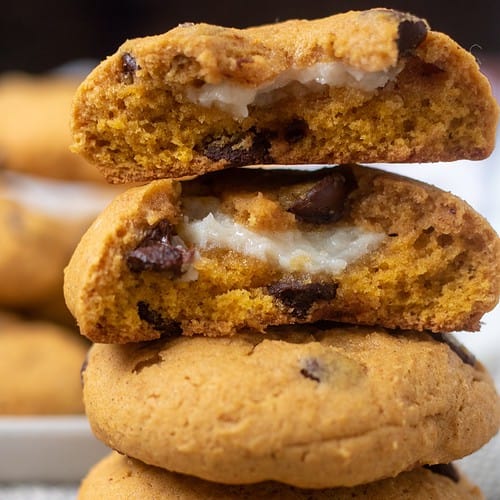 A stack of pumpkin chocolate chip cookies where the top cookie is in half and you can see the cream cheese filling. You can see a beige plate of cookies in the background