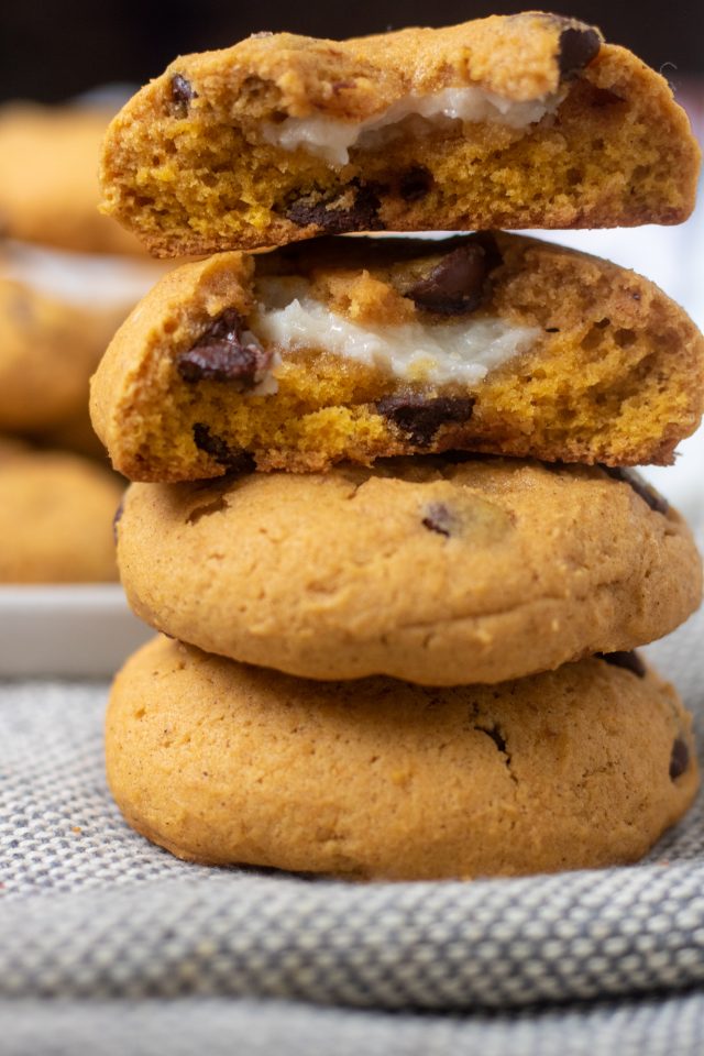 A stack of pumpkin chocolate chip cookies where the top cookie is in half and you can see the cream cheese filling. You can see a beige plate of cookies in the background