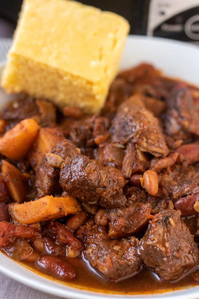 A close up show of chunk beef chili made with chuck roast, sweet potatoes, and beans. A slice of cornbread is on the edge of the bowl.