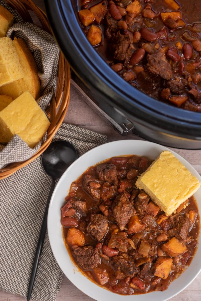 An overhead shot of a white bowl of chili with a side of cornbread, a black crockpot and a basket full of cornbread. There's also a black spoon next to the bowl of chili