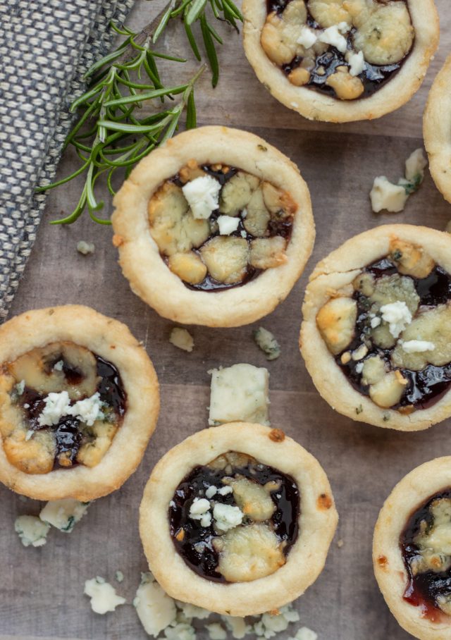 Mini tarts topped with gorgonzola cheese and filled with blackberry jam. There's gorgonzola crumbles sprinkled on a wood background with a fresh rosemary sprig.
