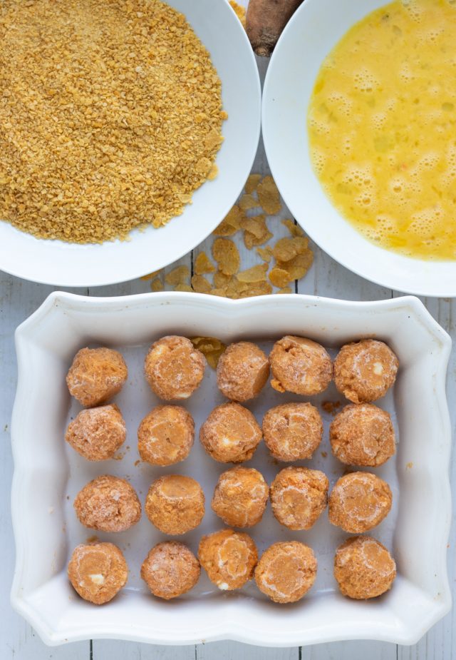 A rectangle white baking dish filled with sweet potato & goat cheese balls. There's a white bowl with corn flake crumbs and a bowl with beaten eggs.