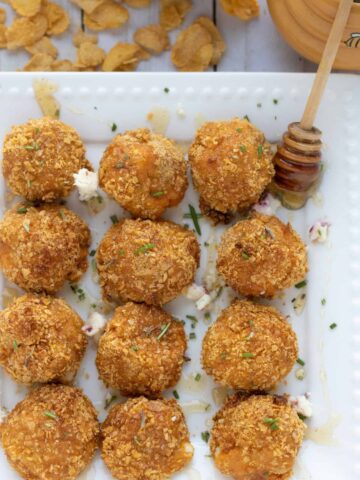 A square white dish with air fried goat cheese and sweet potato balls. They're drizzled with honey. you can see the honey stick with honey oozing off of it. The dish is sprinkled with fresh rosemary and there's corn flakes in the background